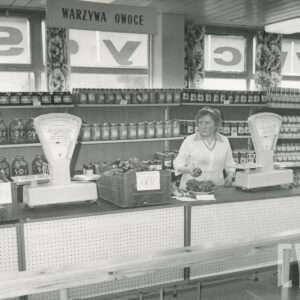 wnętrze sklepu, za długą ladą z wagami i skrzynkami z towarem stoi kobieta w jasnym fartuchu, za nią, w głębi półki ze słoikami