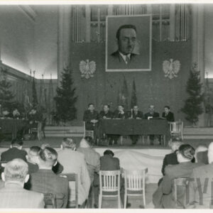 wnętrze dużej sali, na podwyższeniu stół zakryty ciemnym suknem, przy nim siedzi sześciu mężczyzn, za nimi w głębi wisi duży portret mężczyzny, po jego bokach orły, z lewej na mównicy przemawia mężczyzna, na pierwszym planie w rzędach siedzą mężczyźni