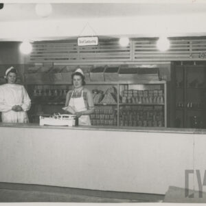 za ladą stoją dwie kobiety, na ladzie stoi waga szalkowa, za nimi półki z towarem