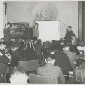 sala wykładowa, na krzesłach, tyłem siedzą w większości mężczyźni, w głębi z lewej tablica, przy niej dwie kobiety, nad nimi na ścianie wisi krzyż z figurą, z prawej stoi mężczyzna