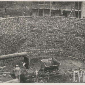 wnętrze budowanego pawilonu, fragment ceglanej ściany, za nią drewniane rusztowania, pracujący robotnicy, na pierwszym planie wagonik na szynach