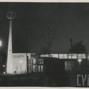 sceneria nocna, murowany budynek z przeszkleniami i półokrągłym wejściem, przed nim spacerujący ludzie, przy budynku murowana kolumna o kwadratowej podstawie, zwężająca się ku górze, na jej wierzchołku przenikające obręcze, na ścianie napis „Społem”