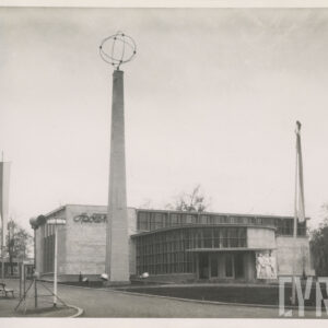 murowany budynek z przeszkleniami i półokrągłym wejściem, przed nim spacerujący ludzie, przy budynku murowana kolumna o kwadratowej podstawie, zwężająca się ku górze, na jej wierzchołku przenikające obręcze, na ścianie napis „Społem”