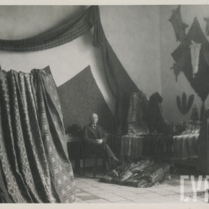 stoisko z tkaninami i futrami, na krześle, w głębi siedzi mężczyzna, z prawej stoi mężczyzna w płaszczu