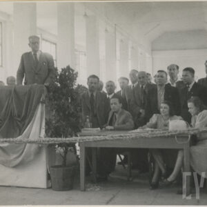 duża hala, z lewej mównica, przy niej stoi mężczyzna, za nim duża grupa ludzi