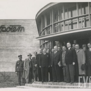 grupa mężczyzn pozuje do zdjęcia na tle półokrągłego budynku, z lewej, w głębi ściana z napisem „Społem”