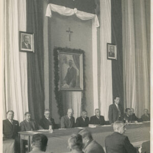 duża sala pod ścianą ustrojoną tkaninami w barwach polskiej flagi, polskim godłem i portretami długi stół, przy nim siedzą mężczyźni, przodem do nich a tyłem do zdjęcia siedzi kilku mężczyzn