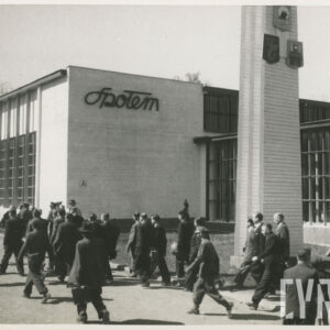 duża grupa ludzi idzie przed murowanym budynkiem z przeszkleniami, przy budynku murowana kolumna o kwadratowej podstawie, zwężająca się ku górze, na ścianie napis „Społem”