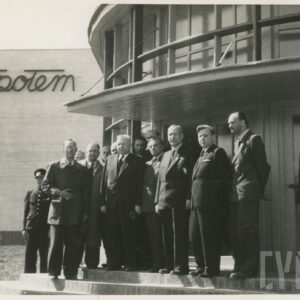 grupa ludzi pozuje do zdjęcia przed murowanym budynkiem z przeszkleniami i półokrągłym wejściem, w głębi na ścianie napis „Społem”