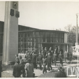 duża grupa ludzi wychodzi z murowanego budynku z półokrągłym wejściem i przeszkleniami, przy budynku murowana kolumna o kwadratowej podstawie, zwężająca się ku górze