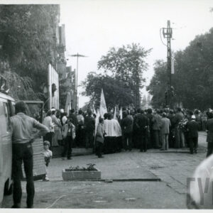 tłum ludzi zebranych na ulicy przy budynku; stoją wśród nich duchowni; część z nich trzyma kwiaty, część sztandary