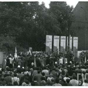 tłum ludzi zebranych na ulicy przy budynku; stoją wśród nich duchowni; część z nich trzyma kwiaty, część sztandary; w głębi widoczne transparenty