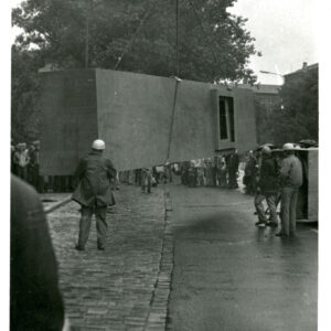 stalowy element wiszący na linach zamontowanych na haku dźwiga; wokół tłum gapiów