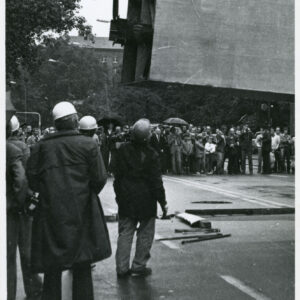 stalowy element wiszący na linach zamontowanych na haku dźwiga; wokół tłum gapiów
