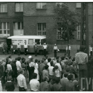 tłum ludzi zebranych na ulicy przy budynku, kilka osób wspięło się na słupy wysokiego napięcia; pod budynkiem stoją samochody