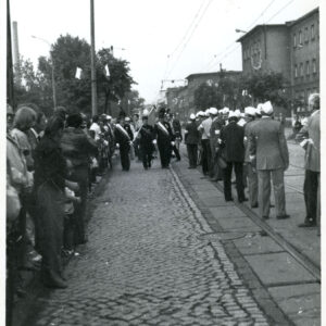 trzech mężczyzn w mundurach górniczych, z górniczymi czapkami niesie sztandar wśród grupy ludzi ustawionej w dwóch rzędach przy ulicy
