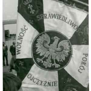 sztandar w biało-czerwoną szachownicę, na środku wyhaftowany biały orzeł, wokół którego wyszyto napis "Wolność, sprawiedliwość, pokój Ojczyźnie"