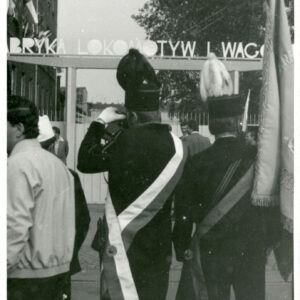 grupa ludzi ludzi zebranych przy bramie; stoi wśród nich dwóch górników w górniczych mundurach z czapkami