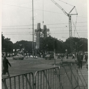 stalowe krzyże otoczone rusztowaniem przy skrzyżowaniu ulic, po których jeżdżą samochody, w tle przechodzą piesi