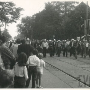 tłum maszerujący ulicą, wzdłuż której rosną drzewa i stoją ceglane budynki; lampy uliczne mają zamontowane nagłośnienie i przystrojone są biało-czerwonymi flagami