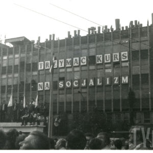 budynek fabryczny z zamontowanymi literami tworzącymi napis: "trzymać kurs na socjalizm", pod budynkiem stoi duża grupa ludzi, część z nich trzyma w rękach sztandary