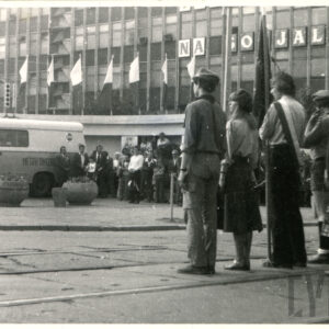 grupa harcerze w poczcie sztandarowym, stojąca przy ulicy; w głębi budynek i samochód