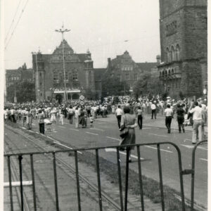 tłum ludzi zgromadzony na ulicy; w głębi reprezentacyjne budynki z cegły, po prawej monumentalny budynek z piaskowca 