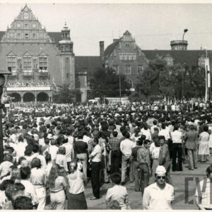 tłum ludzi zgromadzony na ulicy; w głębi reprezentacyjne budynki z cegły, po prawej monumentalny budynek z piaskowca 