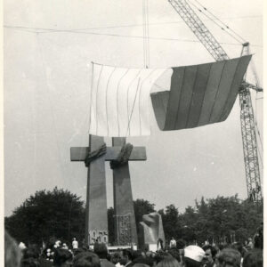 tłum ludzi przed pomnikiem przedstawiającym dwa krzyże; obok dźwig na którego haku przymocowano dwie płachty: białą i czerwoną, symbolizujące flagę Polski