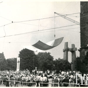 tłum ludzi przed pomnikiem przedstawiającym dwa krzyże; obok dźwig na którego haku przymocowano dwie płachty: białą i czerwoną, symbolizujące flagę Polski