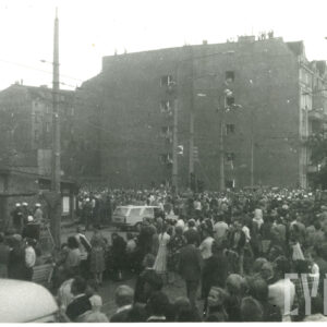 tłum ludzi zgromadzony na ulicy, pomiędzy budynkami mieszkalnymi, na dachu jednej z kamienic stoją ludzie