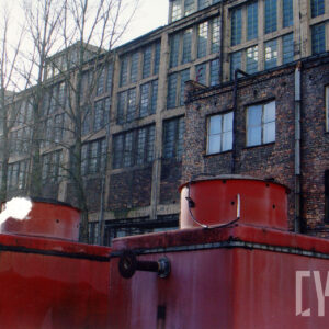 wysoka, długa przeszklona hala warsztatowa, przed nią dobudowany niższy, ceglany budynek, na pierwszym planie dwa czerwone, metalowe zbiorniki