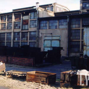 budynek warsztatów z dużymi oknami, i metalowymi wysokimi drzwiami, w części okien wybite szyby, przed budynkiem metalowe pojemniki z gruzem