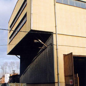 wysoka hala, z wysuniętą górną częścią, w wyższej partii na ścianie szczytowej dwa rzędy okien, u dołu z prawej otwarta metalowa brama, pod halą tory kolejowe, na nich wagonik typu platforma