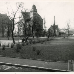 plac z trawnikiem, krzewami i drzewami, w głębi kilkukondygnacyjne budynki z bogatą dekoracją architektoniczną