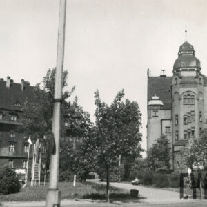 plac z trawnikiem, krzewami i drzewami, w głębi kilkukondygnacyjne budynki z bogatą dekoracją architektoniczną