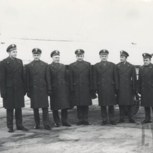 siedmiu mężczyzn stoi w jednym szeregu w mundurach i czapkach wojskowych, pozują na tle słabo widocznego samolotu myśliwskiego