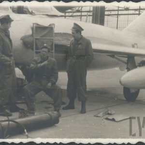trzech mężczyzn w mundurach, mężczyzna w środku siedzi, za nimi fragment samolotu wojskowego