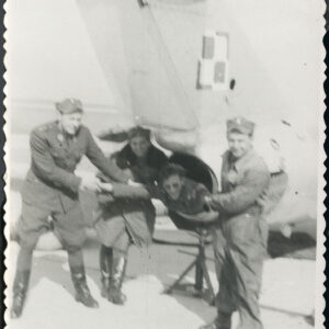 trzech mężczyzn w mundurach stoi przy samolocie wojskowym, widoczny jego tylny fragment, w środku mężczyzna w zabawnej pozie pozuje w dyszy wylotowej samolotu