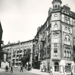 zakrzywiona ulica, przy niej kilkukondygnacyjne kamienice, część z nich z bogatą dekoracją architektoniczną, chodnikiem spacerują ludzie