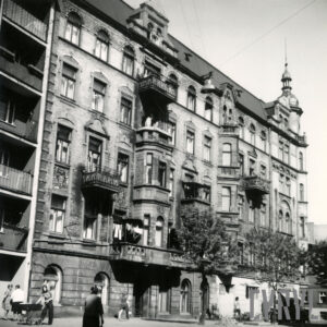 kilkukondygnacyjna kamienica z balkonami, wykuszami, lewy narożnik kamienicy zakończony hełmem, przed nią drzewa