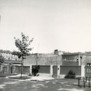 na pierwszym planie płot drewniano-murowany, stojący wzdłuż chodnika, za nim niskie budynki ceglane