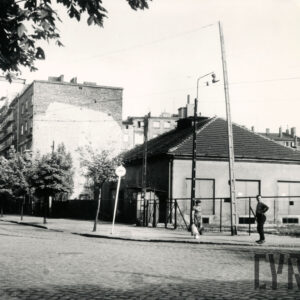 na pierwszym planie brukowana ulica, z prawej niski budynek ze spadzistym dachem, bez okien, za nim kilkukondygnacyjne budynki mieszkalne