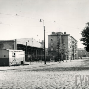 na pierwszym planie brukowana ulica, na ogrodzonym terenie z lewej barak, za nim niska hala z ośmioma otwartymi bramami, w głębi kilkukondygnacyjne budynki mieszkalne