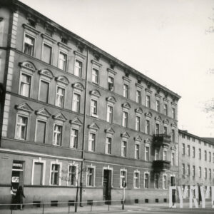 czterokondygnacyjna kamienica narożnikowa, za nią na prawo trzykondygnacyjny budynek, bez zdobień i balkonów