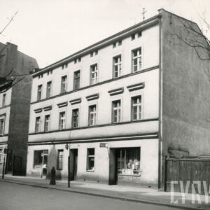 trzykondygnacyjny, prosty w formie budynek, z prawej fragment drewnianego ogrodzenia