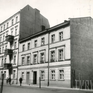 trzykondygnacyjny, prosty w formie budynek, z lewej pięciokondygnacyjna kamienica z balkonami, chodnikiem spacerują ludzie