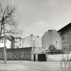 narożnik ulic, na pierwszym planie jednokondygnacyjny, ceglany, prosty budynek, z prawej ogrodzenie, w głębi ściany bez okien wyższych budynków, z lewej latarnia i fragment drzewa bez liści