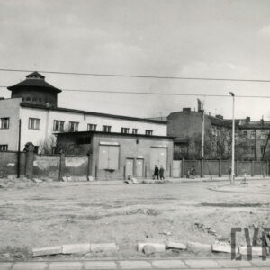 murowany płot, w środku niski budynek bez okien, w głębi kilkukondygnacyjne budynki