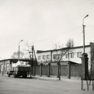 murowany płot, z lewej niski budynek bez okien, w głębi dwukondygnacyjny otynkowany, prosty budynek